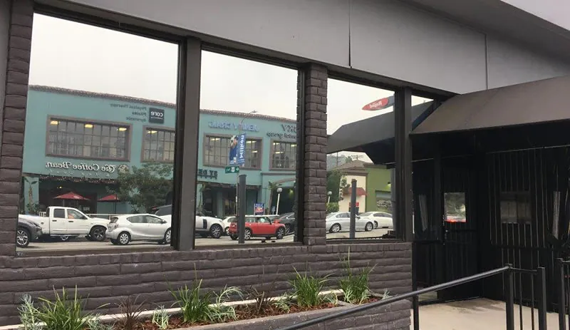 Brick storefront windows with tint film reflecting the shops and parked cars across the street.
